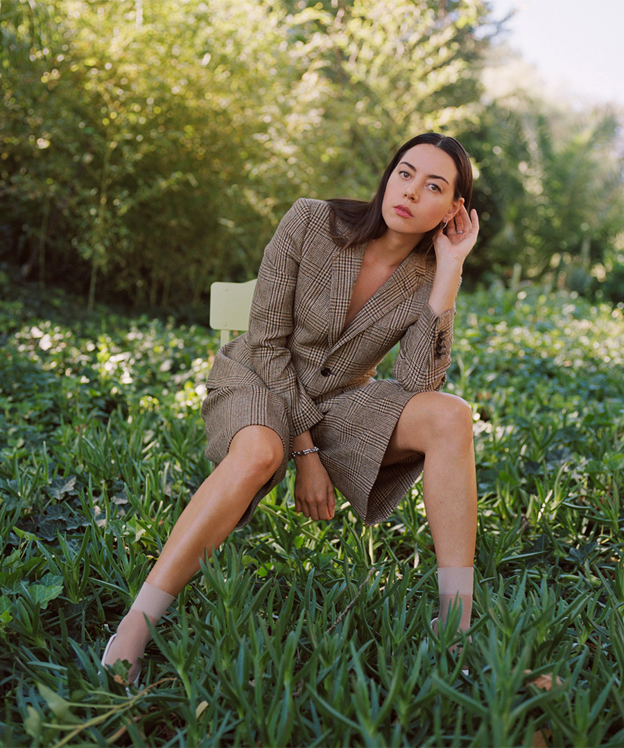 Aubrey Plaza sitting in a green field on a chair, wearing a brown suit.