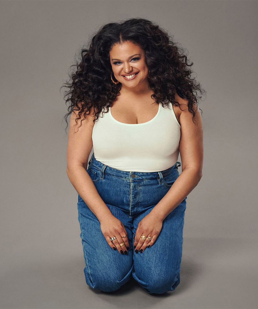 Michelle Buteau in white tank and blue jeans, kneeling with hands on lap.
