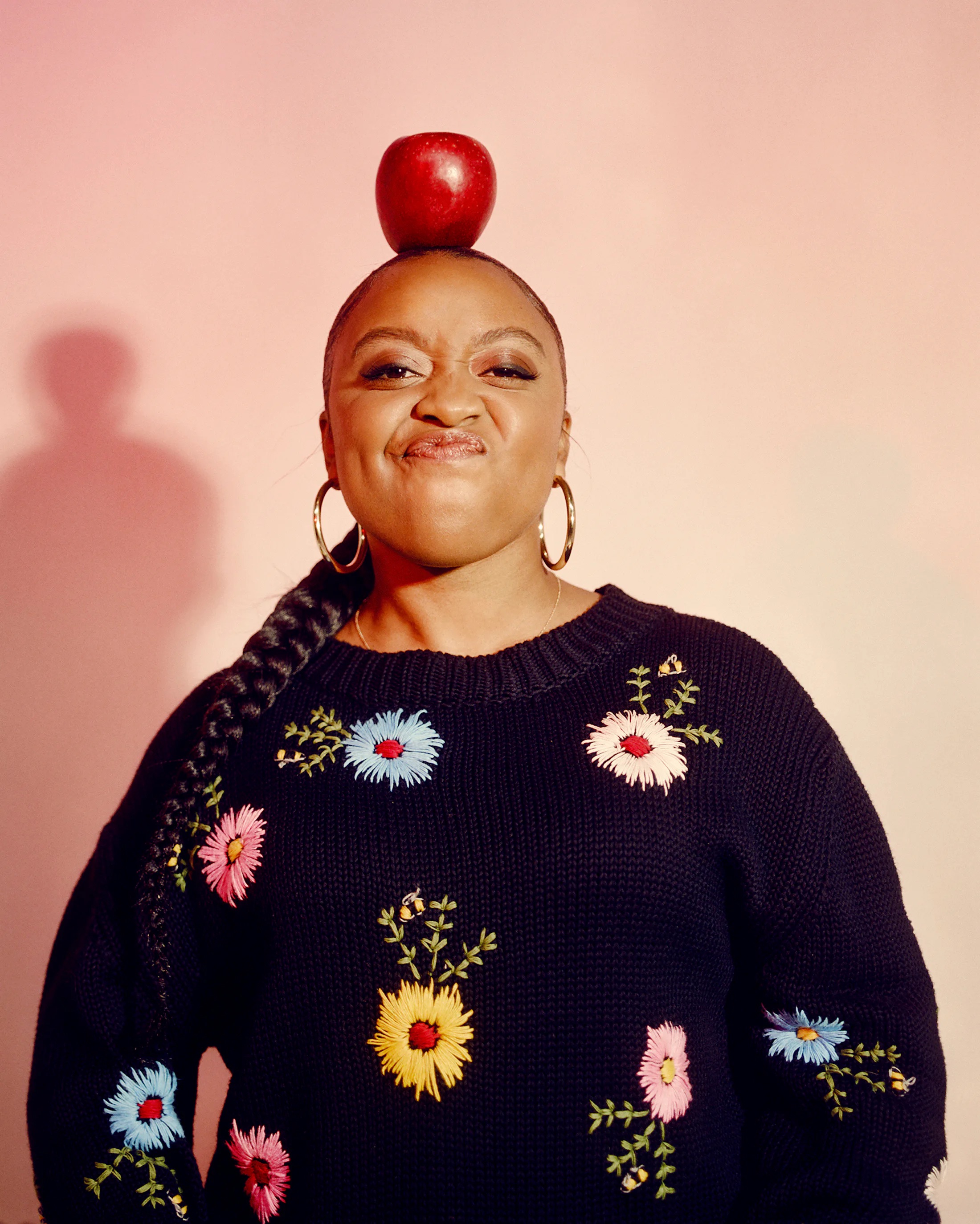Quinta Brunson in black sweater with colorful flowers balancing an apple atop her head against a pink background.