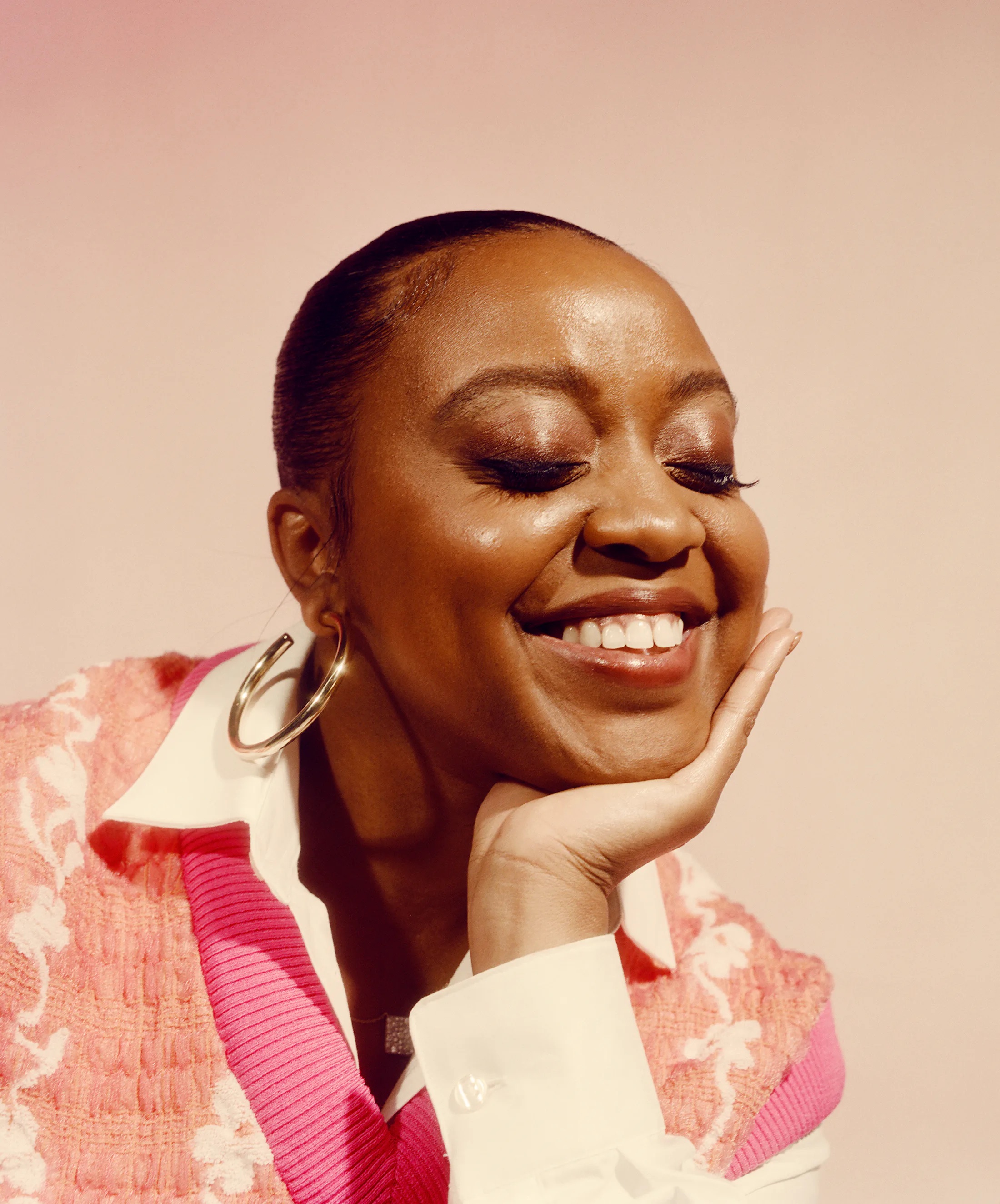 Quinta Brunson with her eyes closed and smiling, resting her chin on her palm in a pink vest and white collar.