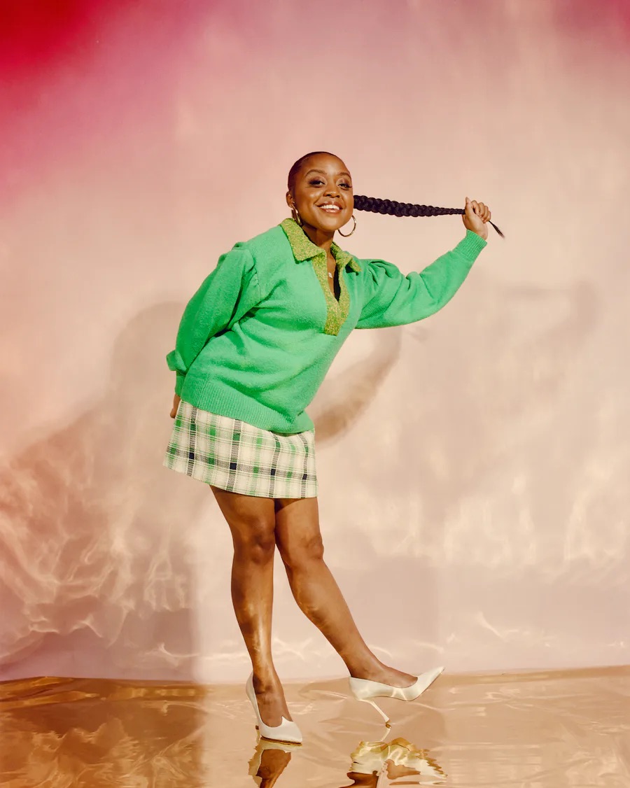 Quinta Brunson in green sweater and plaid skirt holding her ponytail in one hand and in a pose.