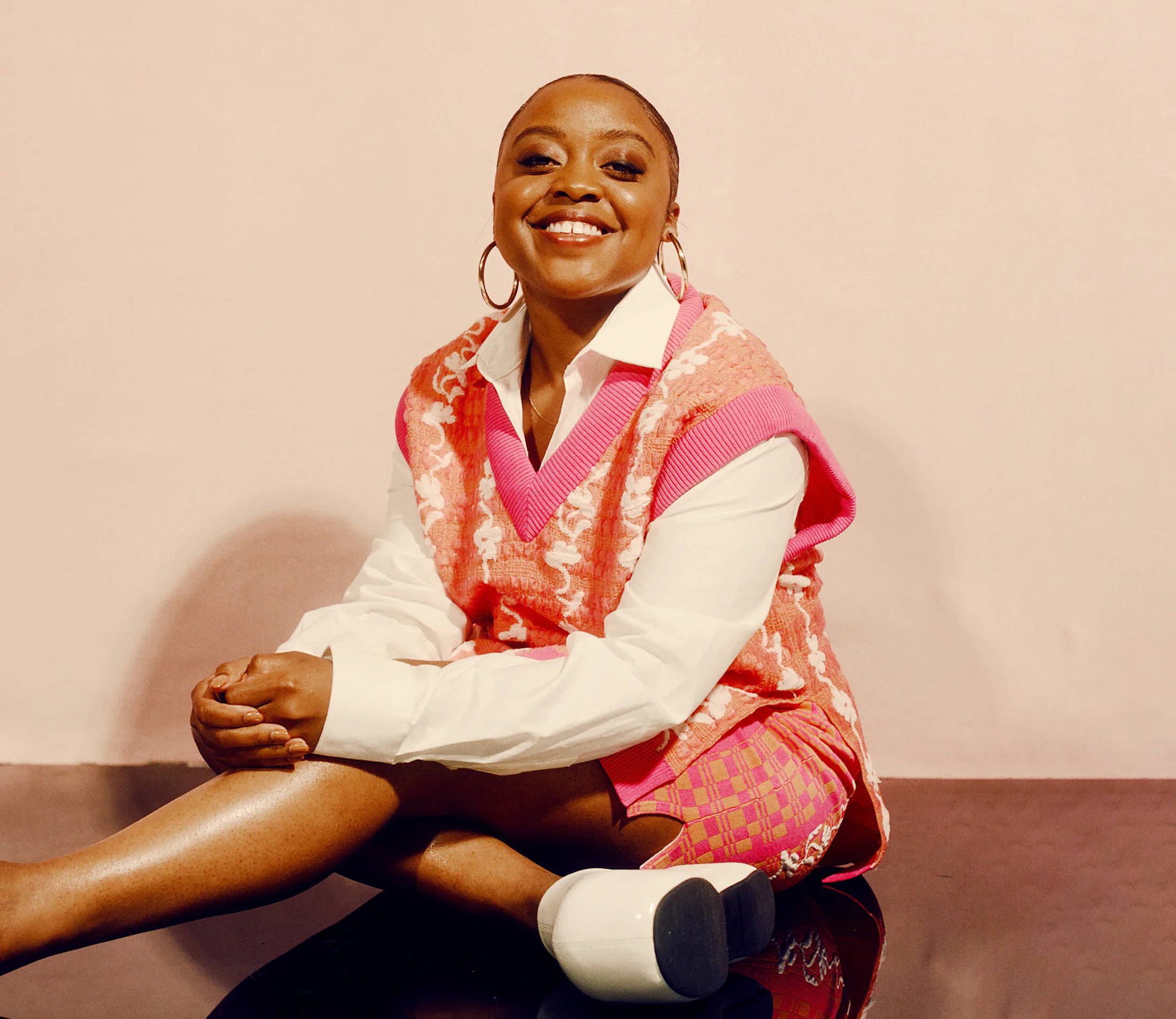 Quinta Brunson in pink vest over white top sitting down cross-legged against a light peach background.